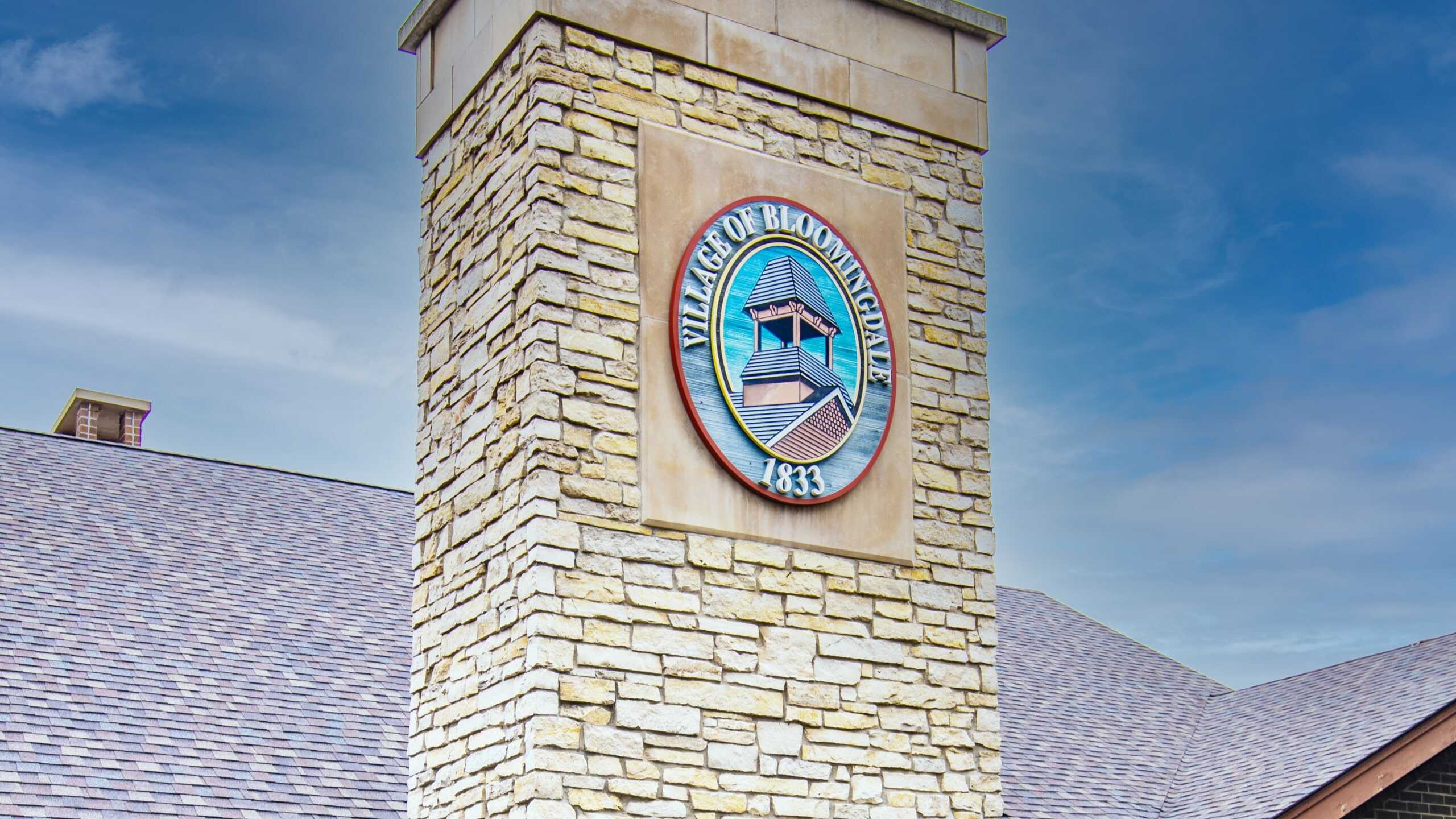 Stone tower featuring the Village of Bloomingdale, Illinois emblem under a blue sky.